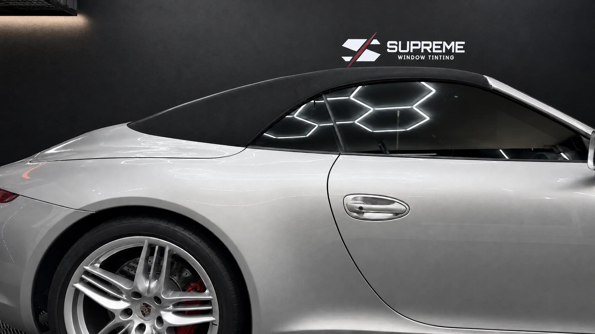 Silver Porsche 911 Cabriolet with freshly tinted windows under hexagon lights in the Supreme Window Tinting garage