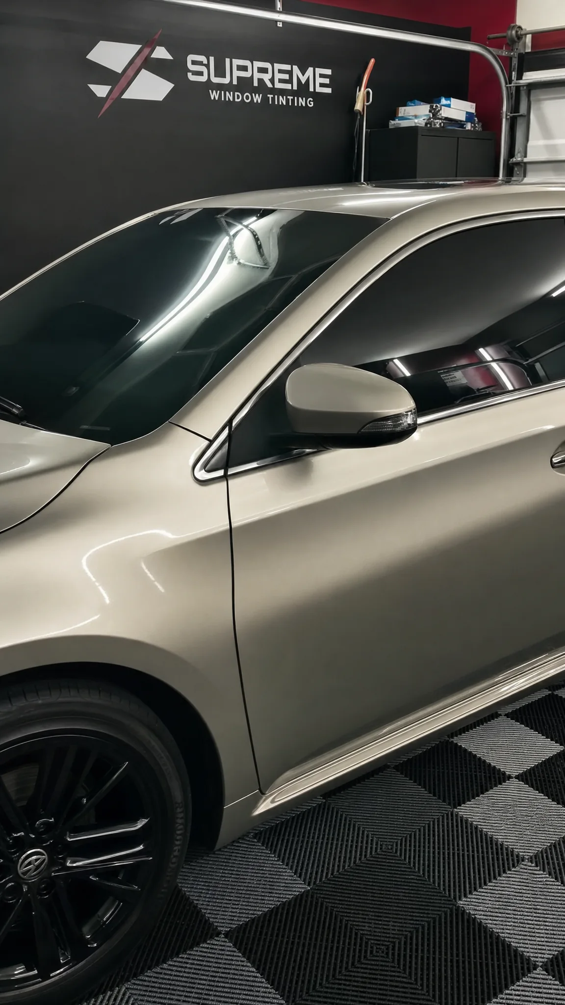 Tinted side glass on a gray sedan, indoors with the Supreme Window Tinting logo on the wall